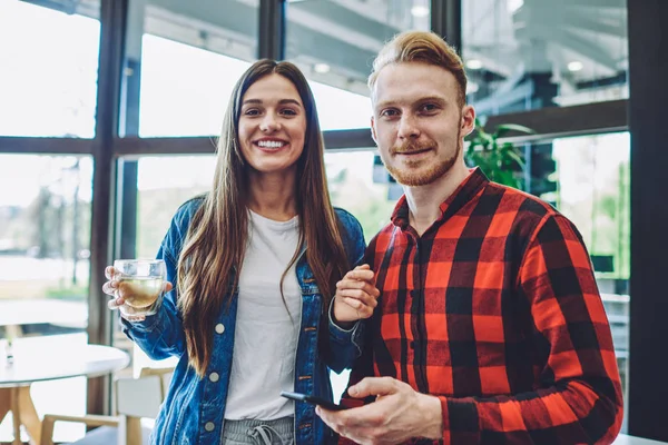 Positive millennial man and woman looking at camera while spending time in cafeteria for drinking taste beverage and using free internet connection on smartphone gadget for communicate online