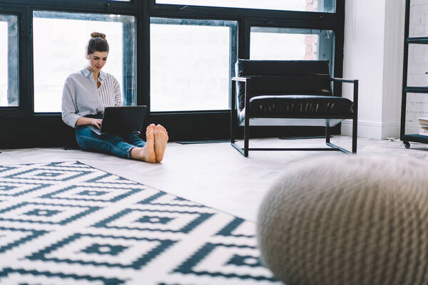 Caucasian young woman sitting on floor and working remotely at modern laptop using wireless internet connection in stylish apartment, skilled hipster girl searching information and installing app