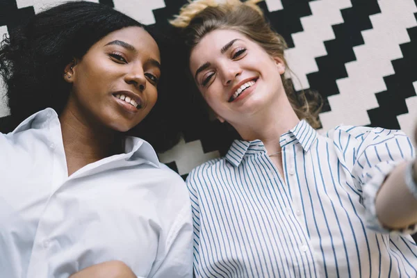 Top view of positive multicultural hipster girls smiling at camera while lying on apartment floor, portrait of happy two diversity young women taking rest at home and spending time together