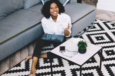 Mutlu Afro-Amerikan hipster kız portresi kahve masasının yanında oturuyor ve boş zamanlarını çay için harcayıp internetten modern dokunmatik ped üzerinden sohbet etmekten zevk alıyor.