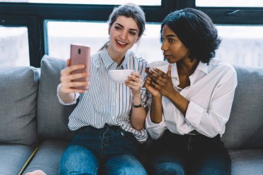 Çekici siyah tenli kız ve akıllı telefonun kamerasında poz veren neşeli beyaz arkadaş sosyal ağlarda yayınlanmak için güzel selfie çekiyorlar, hücrede mutlu kadınlar fotoğraf çekiyor.