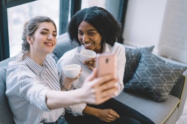 Mutlu iki bayan arkadaş dijital akıllı telefonun önünde gülümsüyor ve fotoğraf çekiyor, neşeli çok kültürlü kızlar evde cep telefonuyla selfie çekiyorlar kahve sohbetleri sırasında.