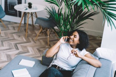 Modern apartmandaki koltukta oturmuş akıllı telefondan gülümseyen ve konuşan neşeli afro-amerikalı hippi kız dairedeki cep telefonundan arıyor.