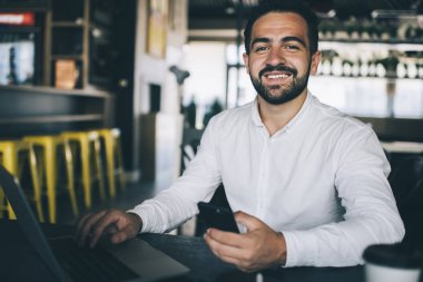 Gündelik beyaz gömlekli, cep telefonu cihazıyla gezen ve kameraya gülümseyen neşeli erkek girişimci portresi. Modern dizüstü bilgisayarda uzaktan iş yapan mutlu adam dijital teknolojide 4G kullanıyor.