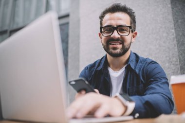 Klasik görüş düzeltmesi için gözlüklü neşeli bir adamın portresi kameraya bakıp gülümserken modern cep telefonu ve laptop cihazları kullanıyor.