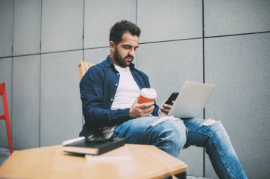 Millennial hipster 'lı adam, modern dizüstü bilgisayarla sokak kafeteryasında akıllı telefondan yetenekli erkek mesajlaşması geliştirmek için otururken internetten cep telefonu uyarısını kontrol ediyor.