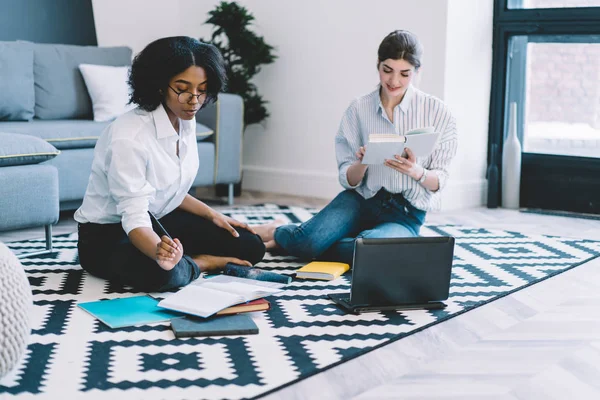 Afro american and caucasian female students having meeting at home apartment for making productive preparation to university exam sitting on floor with laptop computer and other equipment for studying