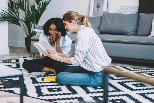 Amazed multicultural women shocked from read information from literature book sitting on floor in apartment with stylish interior, happy hipster girls surprised from organisation plan in textbook