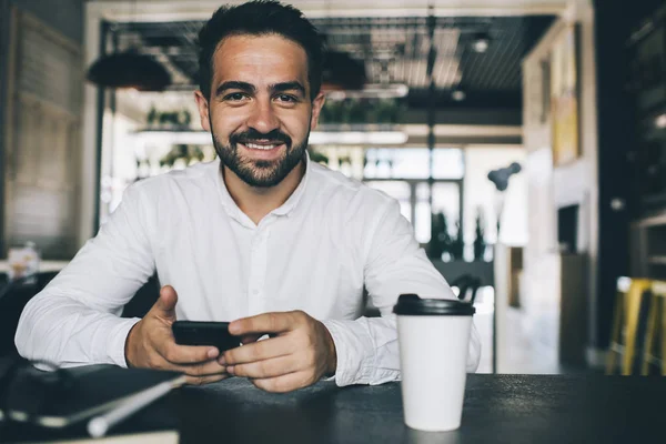 Portrait of positive successful male entrepreneur with mobile phone in hands spending work break in coffee shop sitting at cafeteria table with takeaway caffeine beverage and smiling at camera