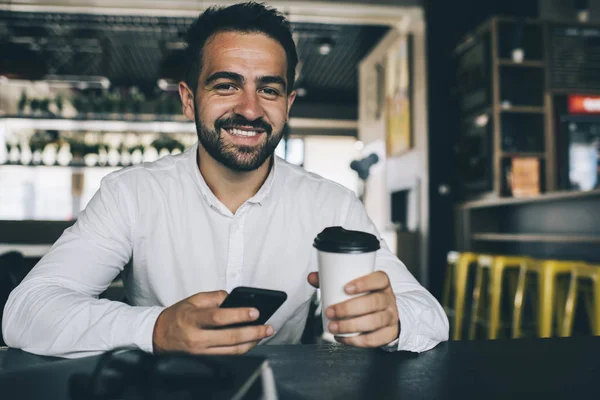 Portrait of happy accomplished male manager of coffee shop holding cell smartphone gadget with 4g wireless for browsing websites and smiling at camera during digital break, experienced communication