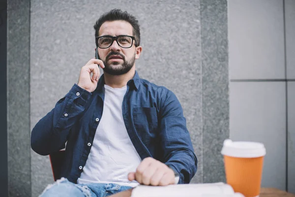 Thoughtful hipster guy dressed in casual wear making online intrenational cell call for talking and consultancy with operator about 4g roaming internet, concept of technology and communication