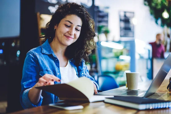 Happy female student analyzing information from knowledge textbook during exam preparation in university cafeteria while sitting at table with modern laptop computer, positive woman studying