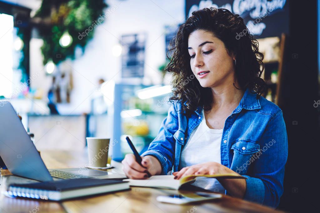 Concentrated female author writing plot for next bestseller in textbook with interesting idea, caucasian woman in denim t shirt making notes for organisation plan during learning in cafe interior