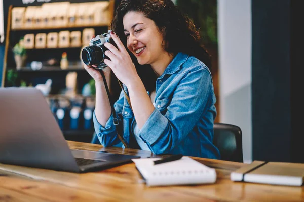 カフェのインテリアで長距離の仕事のための時間の間にコンピュータの画面の写真を撮る中で良い感じの幸せな女性写真家 古いスタイルのカメラを使用して正の白人のヒップスター女の子 — ストック写真