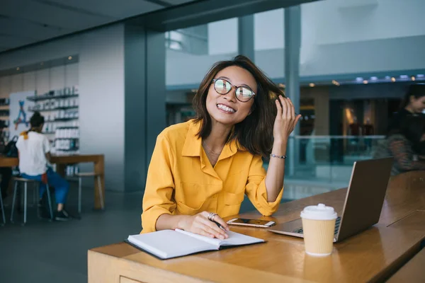 Happy Chinese female student in optical glasses for vision correction rejoicing in coworking space during exam preparation with modern laptop computer and textbook for education, e learning concept