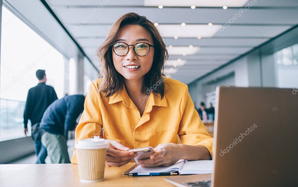 Portrait of Chinese positive hipster girl in trendy spectacles holding cellphone gadget for blogging and smiling at camera during e learning, happy student spending time for knowledge education