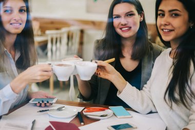 Reklam alanında kahve molası sırasında kameraya bakan ve tezahürat yapan neşeli kadın iş ortaklarının portresi, pozitif gülümseyen kadınların içki bardaklarıyla işbirliği yaptıkları için eğlenmeleri.