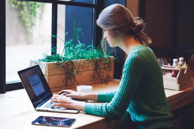 Modern laptop cihazıyla kafeterya masasında otururken ve iletişim için uygulama uygulaması aracılığıyla program kodu yazarken uzaktan çalışan genç bir kadın serbest yazar.