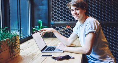 Neşeli hipster adamın portresi dizüstü bilgisayarla oturmuş internet üzerinden alışveriş yapmak için internet bağlantısını bekliyor. Başarılı erkek serbest çalışan kapalı alanda çalışmaktan zevk alıyor.