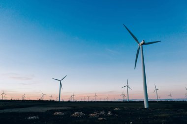 Döner pervaneler ile yel değirmenleri İstasyonu mavi günbatımı gökyüzüne karşı kırsal tarım ortamında Eko kaynaklardan alternatif temiz yeşil güç üreten. Uzak, elektrik üreten türbinlerin