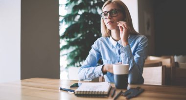 Not defteri ve kalemler ile ahşap masada otururken tasarım projesi için fikirler hakkında düşünme ciddi kadın öğrenci, kampüste gözler koruma öğrenme sağlamak için gözlükler içinde dalgın kadın