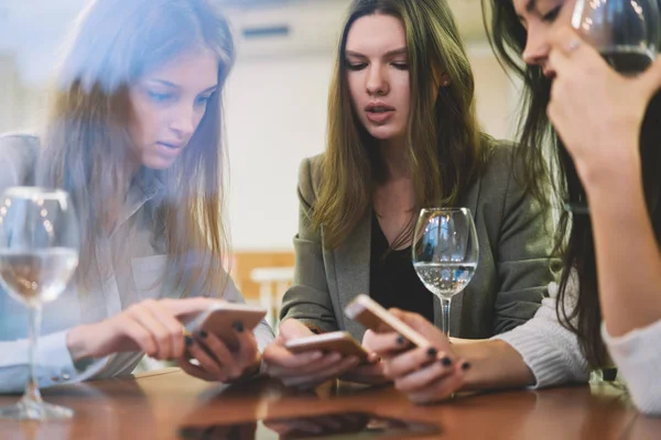 Charming female bloggers checking notification on smartphone gadgets during friendly meeting in cafe interior for drinking white wine, caucasian women reading news in social networks via cellphones