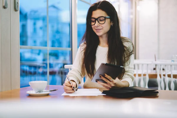 Positive successful hipster girl in optical spectacles for provide eyes protection looking on coffee cup with cute smile on face while holding digital tablet in hand during time for studying