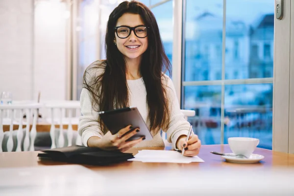 Portrait of cheerful woman in eyewear for vision correction looking at camera during preparing to university examination in coffee shop using 4g internet connection on touch pad for making research