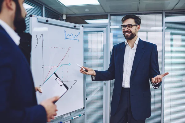 Successful male entrepreneur in optical spectacles for vision correction standing near flip chart with economic graphics and communicating with business partner about main topic of develope company