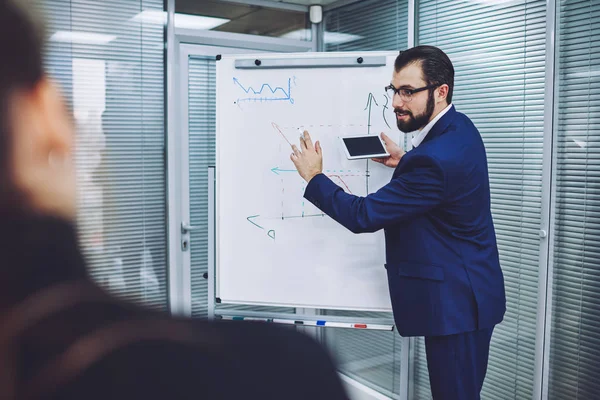 Clever male businessman dressed in elegant formal suit spending time for speaking main information for company workers using graphic on white board and information from mobile phone with mock up