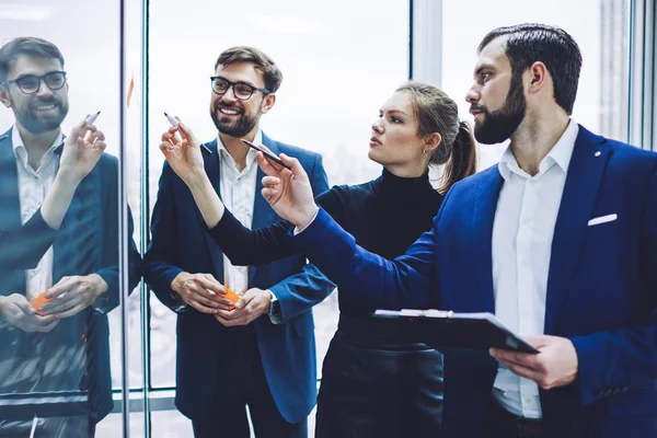 Concentrated colleagues drawing diagram on company window for planning business strategy, collaboration of clever male and female partners near modern board during working process in office