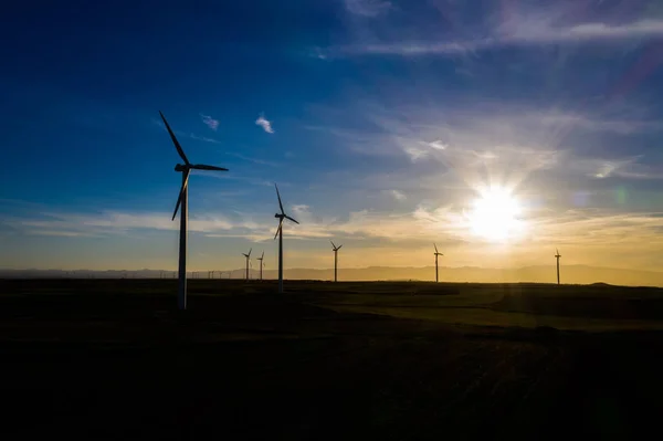 Silhouette Modern windmills with propellers standing in rural farmland agri meadow over orange sunset sky. Wind blowing generating renewable energy for clean resources. Electric power production