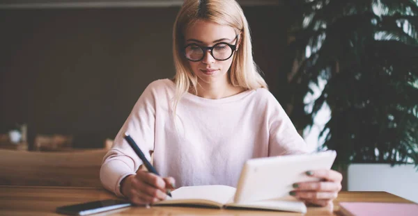 Clever female writer thinking about information for article about millennial people, young caucasian woman in spectacles spending time for studying while sitting at desk with textbook for education