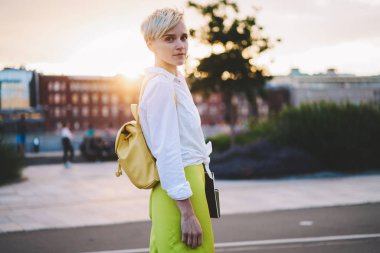 Çekici beyaz hipster kız yarım uzunlukta portre güneş ile kentsel ortamda ayakta sırt çantası ile trendy giyim giyinmiş ve gazeteciler için kurslar için yolda kamera bakıyor