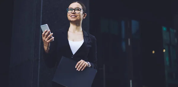 Cheerful woman dressed in formal wear feeling happy from received email with good news about proud ceo using smartphone gadget outdoors, business woman with folder in hand looking away and smiling