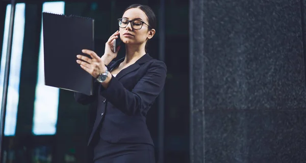 Confident female boss calling to employee for discussing information for presentation in company reading main theses form paper in folder,woman in formal wear concentrated during cellular conversation