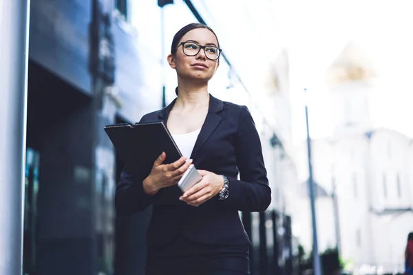 Attractive arrogant female finance director of company looking away during standing on publicity area with paper folder and smartphone gadget in hands, confident economist in optical eyeglasses