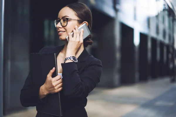 Smiling female finance professional making positive smartphone conversation via roaming connection while spending time on urban setting, happy business woman calling to colleague for consultancy