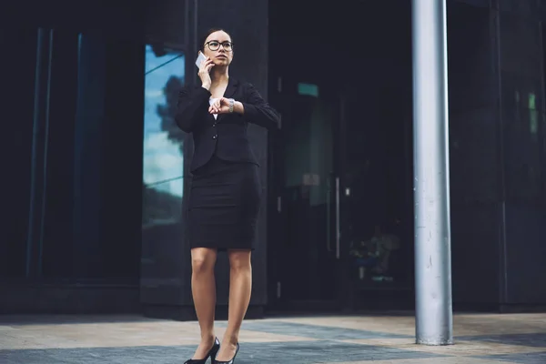都会の設定を待ちながら携帯電話でビジネスパートナーに電話するメガネの真面目な女性 腕時計の時間をチェック スマートフォンで会話をする成功した女性起業家 — ストック写真