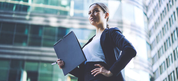 Positive female entrepreneur in formal wear from designer feeling confident and enjoying good day on urban setting, successful caucasian business woman with folder in hand standing on publicity area
