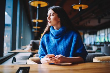 Cep telefonu kullanarak kafeteryaya oturan kadın öğrenci durulmaya ve App online arkadaşlarınızla sohbet, hipster kız akıllı tutarak uzak bakıyor hayal ve arama kapalı bekliyor