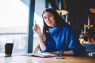 Cafe otururken genç çekici hipster kız gazeteci ve defter yazma olarak çalışan portre, ciddi güzel kadın öğrenci kapalı öğrenme molası kamera bakıyor