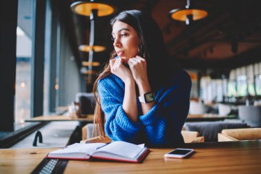 Düşünceli esmer hipster kız rahat kafeteryada arkadaş bekliyor, pencere bakarak dalgın iken, dalgın şık kadın gazeteci ahşap masa kapalı oturma işten mola var