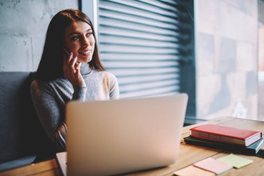 Pencere bakarak ve arkadaş kapalı ile tebliğ için yeni akıllı telefon uygulaması üzerinden arama yaparken kafeterya modern dizüstü bilgisayar kullanarak online çalışan başarılı gülümseyen kadın Yöneticisi