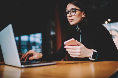 Gelişmiş dizüstü bilgisayar önünde ahşap masada kafede oturan şık gözlükler siyah kazak giymiş genç iş kadını ve el Smartphone tutarken online Webinar izlerken