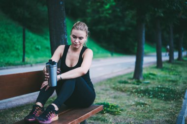 Aktif kadın Runner doğa üzerinde kardiyo eğitim mola zevk alüminyum Tumbler elleri ile ahşap tezgah üzerinde oturan ve germe egzersizleri hakkında düşünme, beyaz kadın açık havada dinlenme
