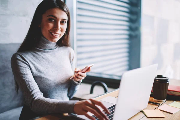 Portrait of successful carefree female blogger using smartphonefor making transaction in web store on laptop computer looking at camera and smiling, millenial hipster girl using gadgets for blogging