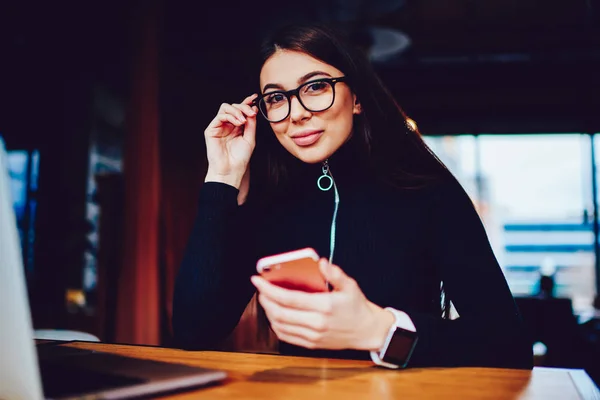 Pozytywna Uśmiechnięta Hipster Dziewczyna Siedzi Kawiarni Patrząc Kamerę Trzymając Smartfon — Zdjęcie stockowe