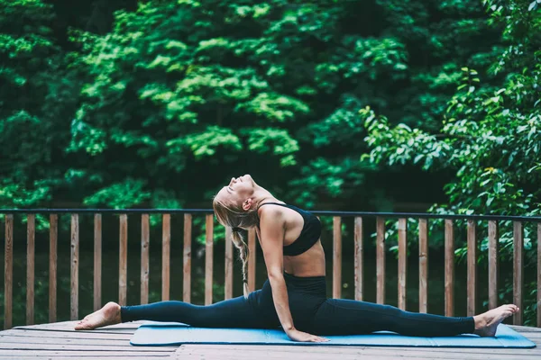 Sportive fitness girl stretching out on caremat and looking up during workout, young woman doing exercises to strengthen her back muscles while practicing yoga and twine on green background