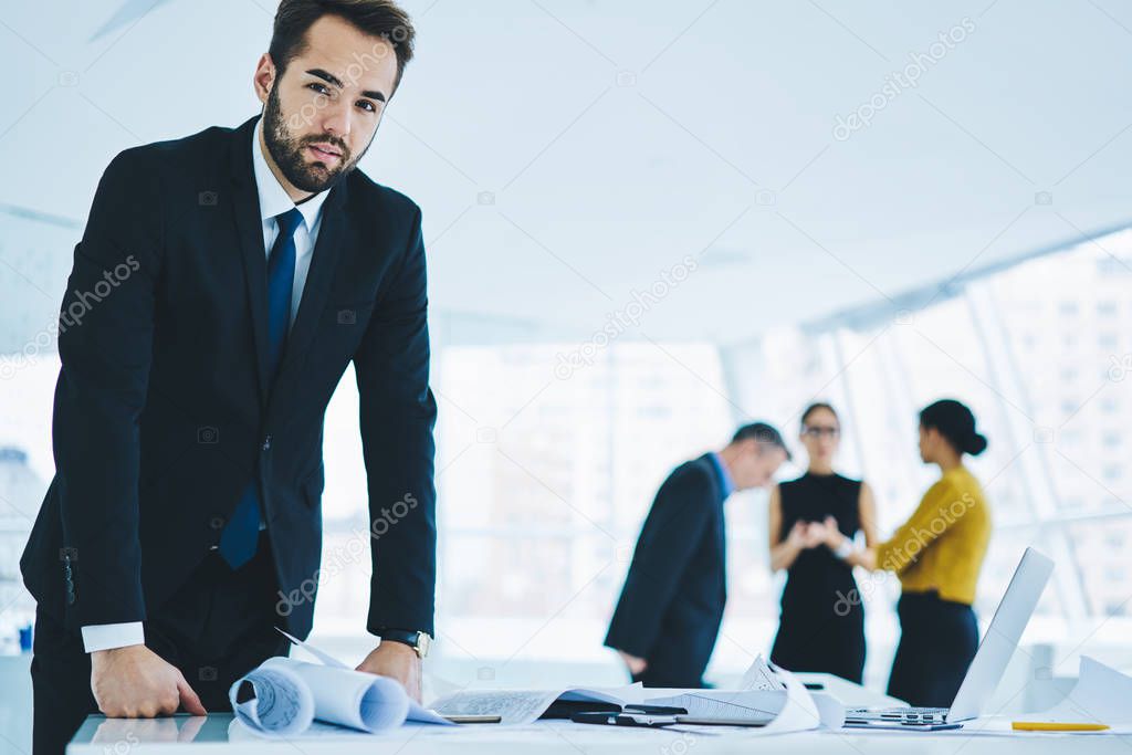 Portrait of mature businessman in formal suit standing near desktop and looking at camera with male and female partners on blurred background, bearded man during working time in corporate company